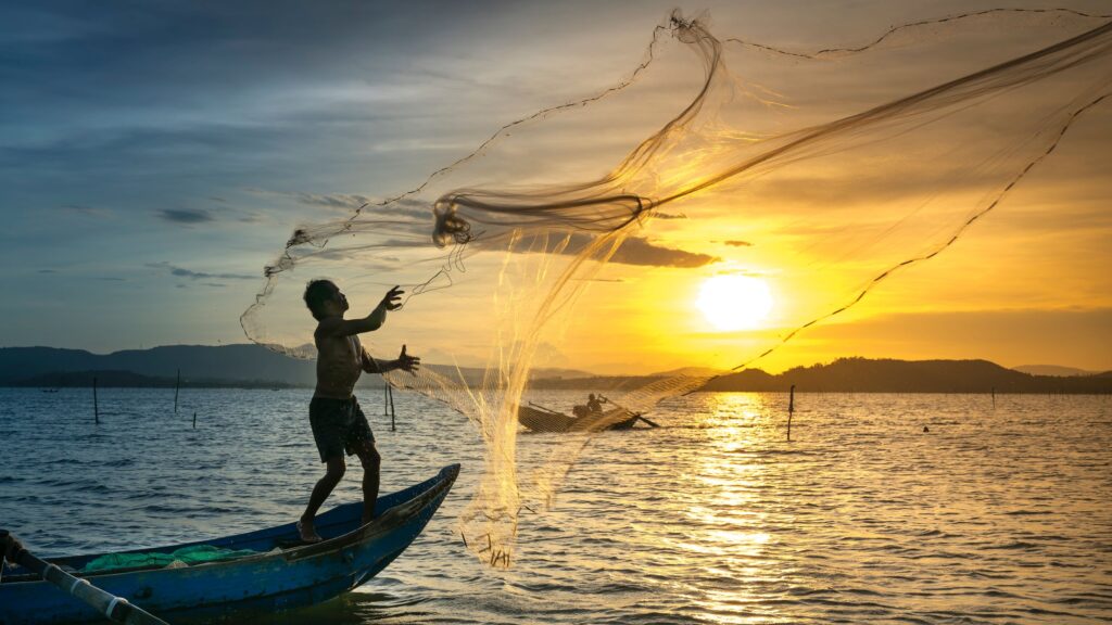 A fisherman stands on a small boat casting a net across a calm lake at sunrise