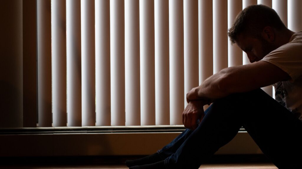 A man sits on the floor beside window blinds with his head resting on his knees