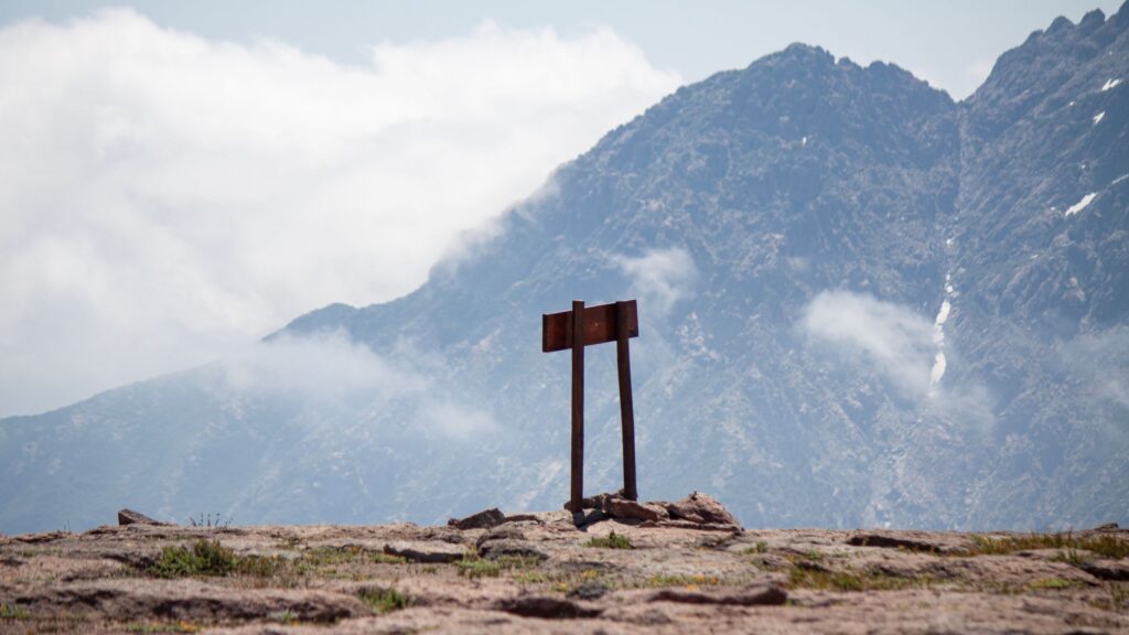 A weathered wooden signpost stands on a rocky ridge with mist-covered mountains behind it